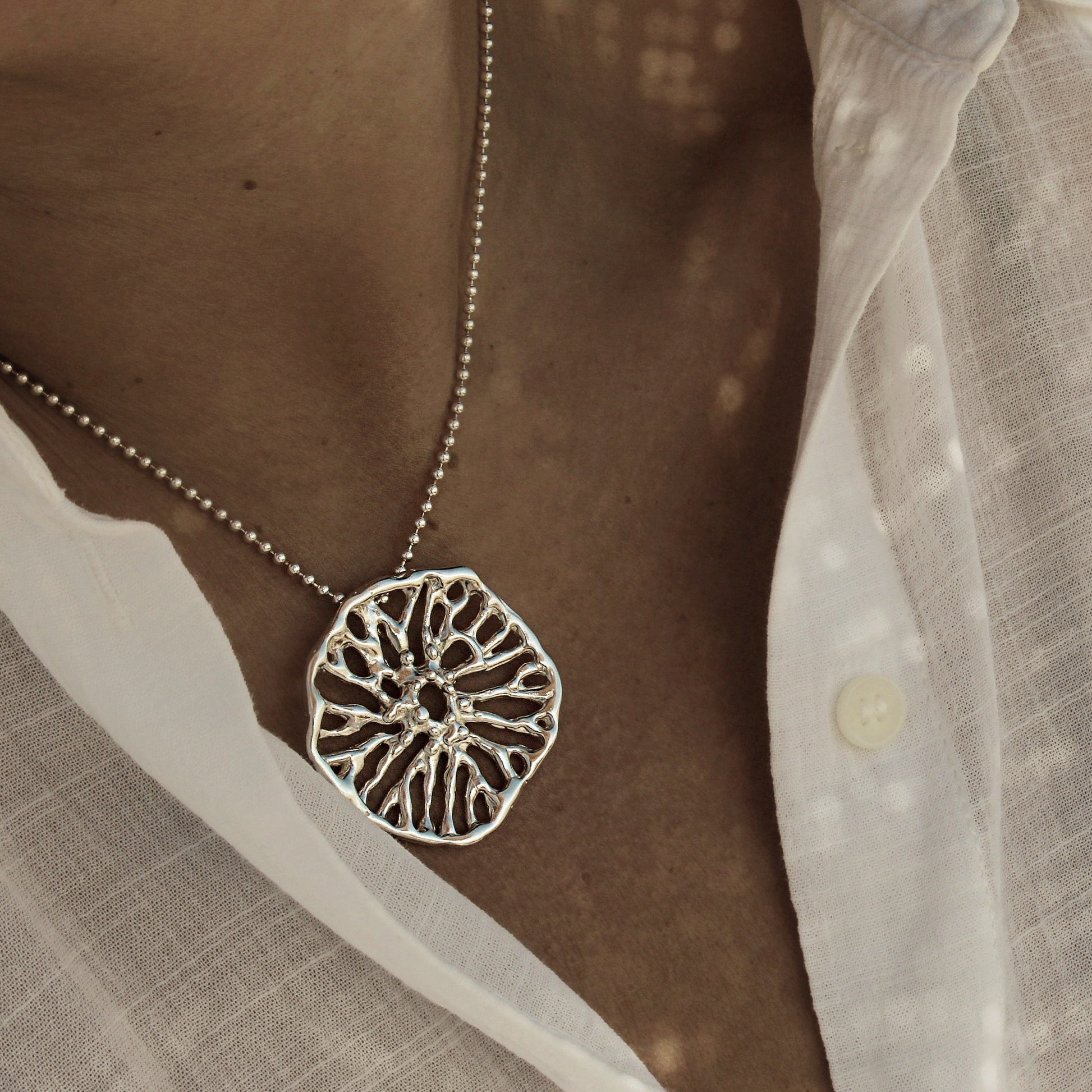 Silver necklace with a detailed pendant worn by a person in a white shirt. Colgante escultórico Flow collar de plata inspirado en el coral hongo, joyería artesanal mediterránea.