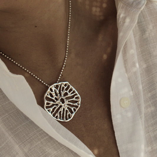 Silver necklace with a detailed pendant worn by a person in a white shirt. Colgante escultórico Flow collar de plata inspirado en el coral hongo, joyería artesanal mediterránea.